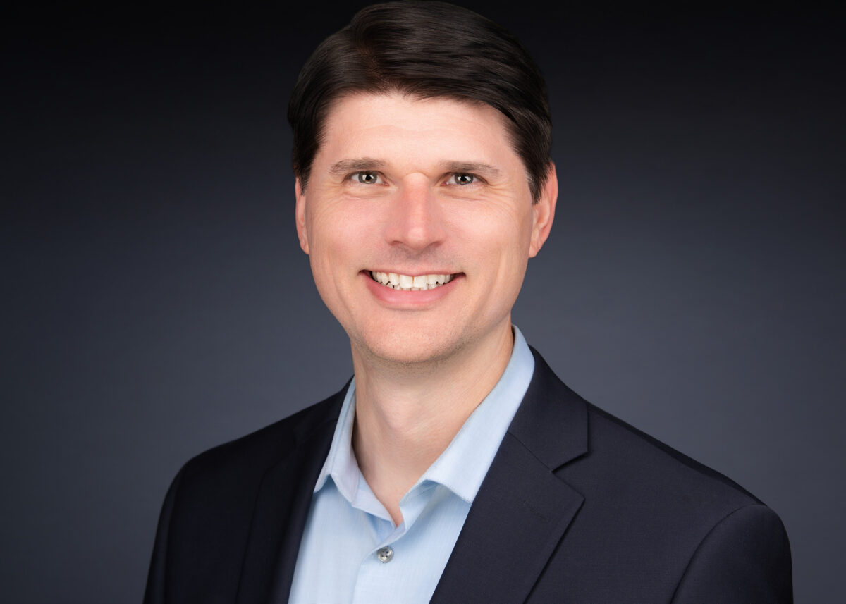 Professional headshot of a man with dark brown hair, wearing a navy blazer over an open-collar light blue shirt, offering a composed, approachable smile against a soft grey gradient background.