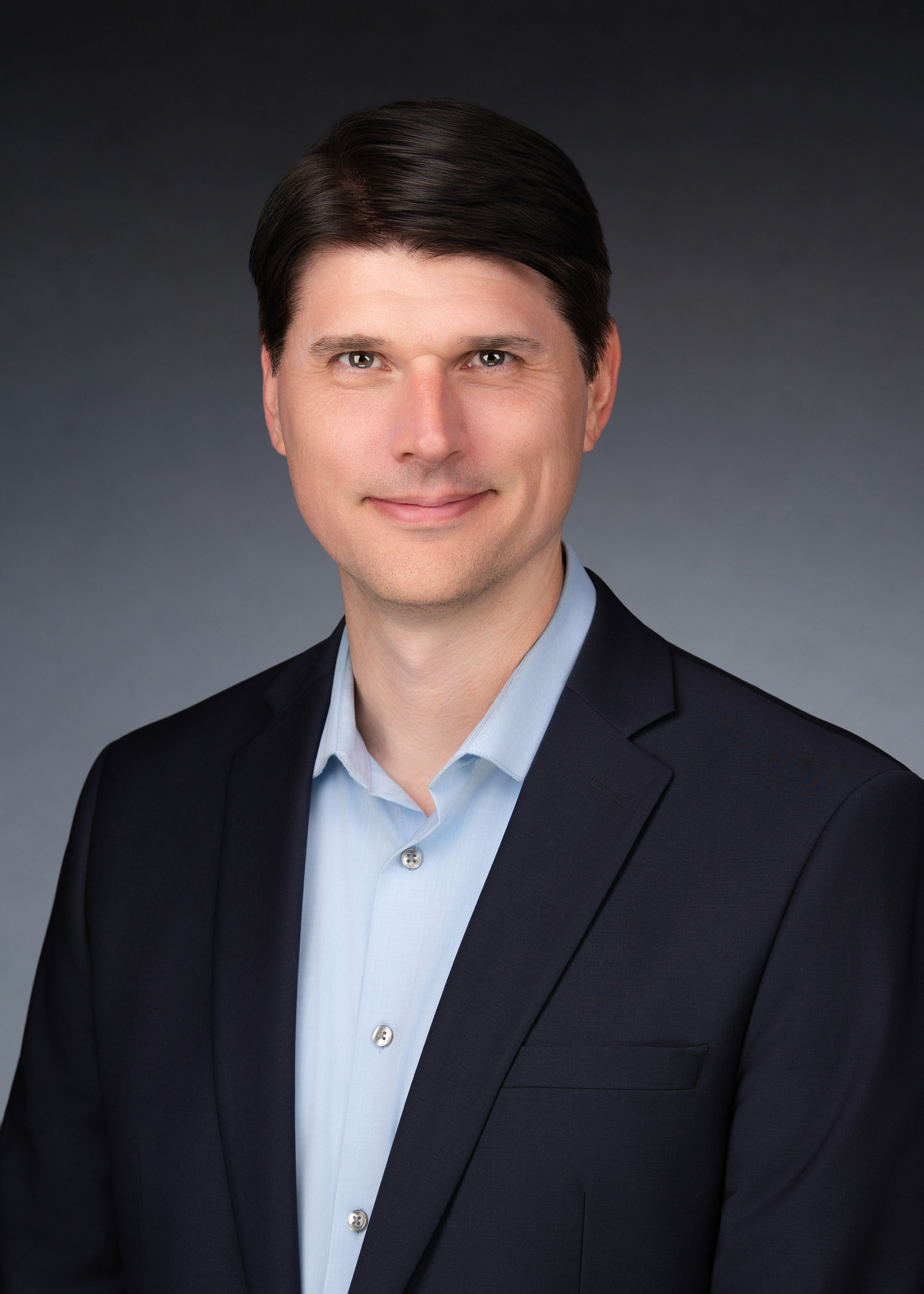 Professional headshot of a man with dark brown hair, wearing a navy blazer over an open-collar light blue shirt, offering a composed, approachable smile against a soft grey gradient background.