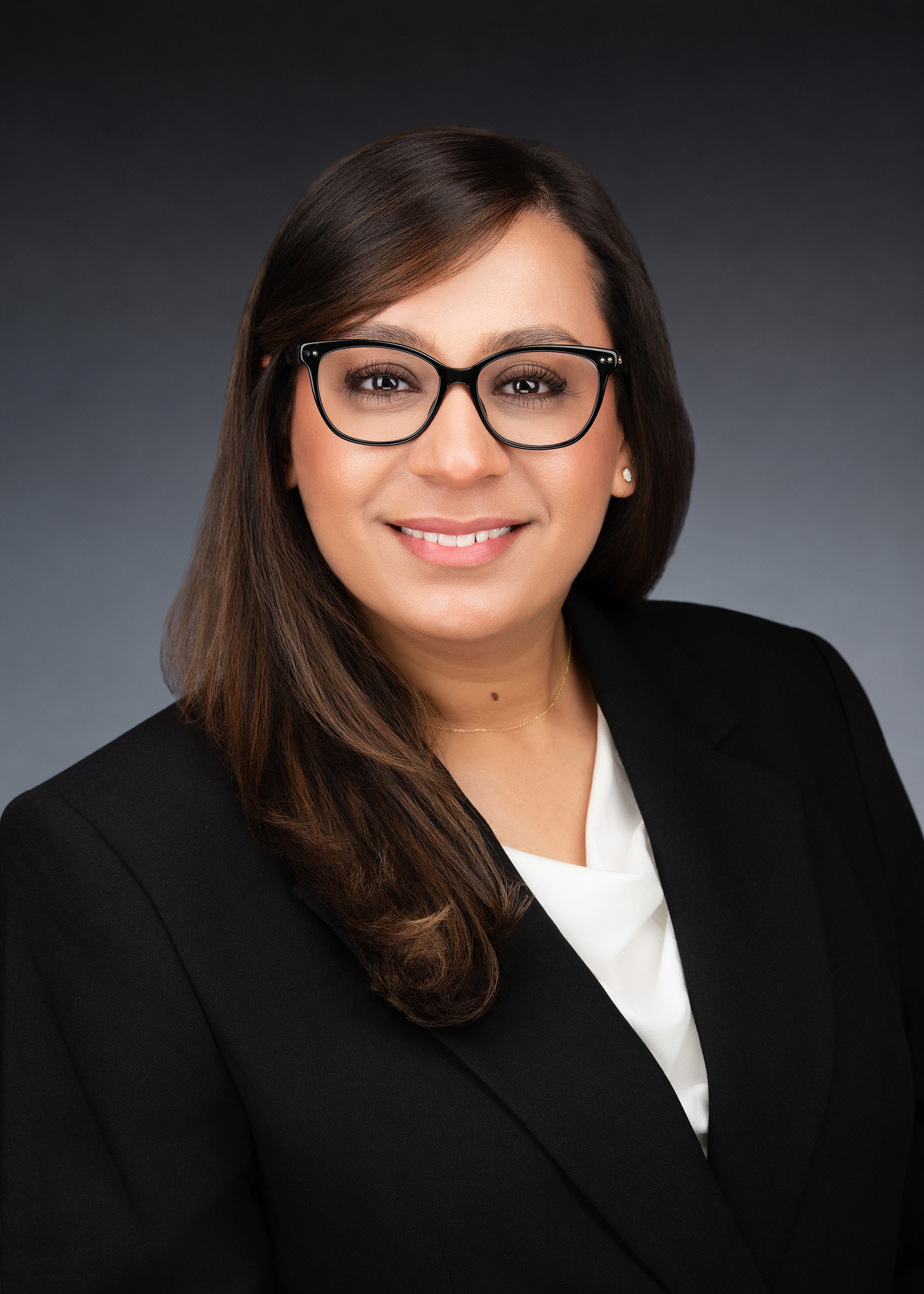 Professional headshot of a woman with long dark brown hair, wearing black-framed glasses and a black blazer over a white blouse, smiling confidently against a soft grey studio background.
