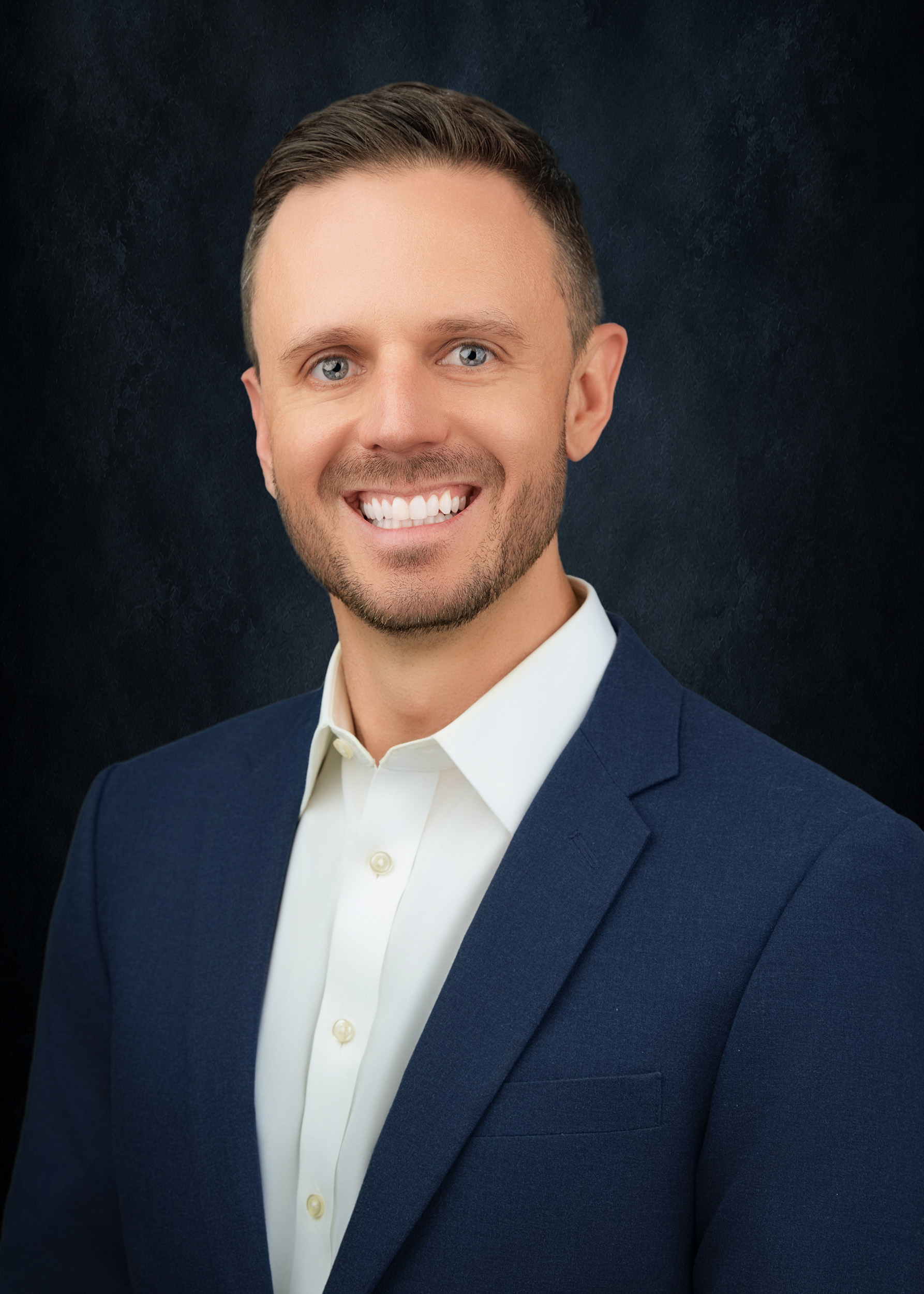 Professional headshot of a man with short dark hair and light blue eyes, wearing a navy blazer over an open-collar cream dress shirt, with light stubble and a broad smile against a textured dark navy studio background.