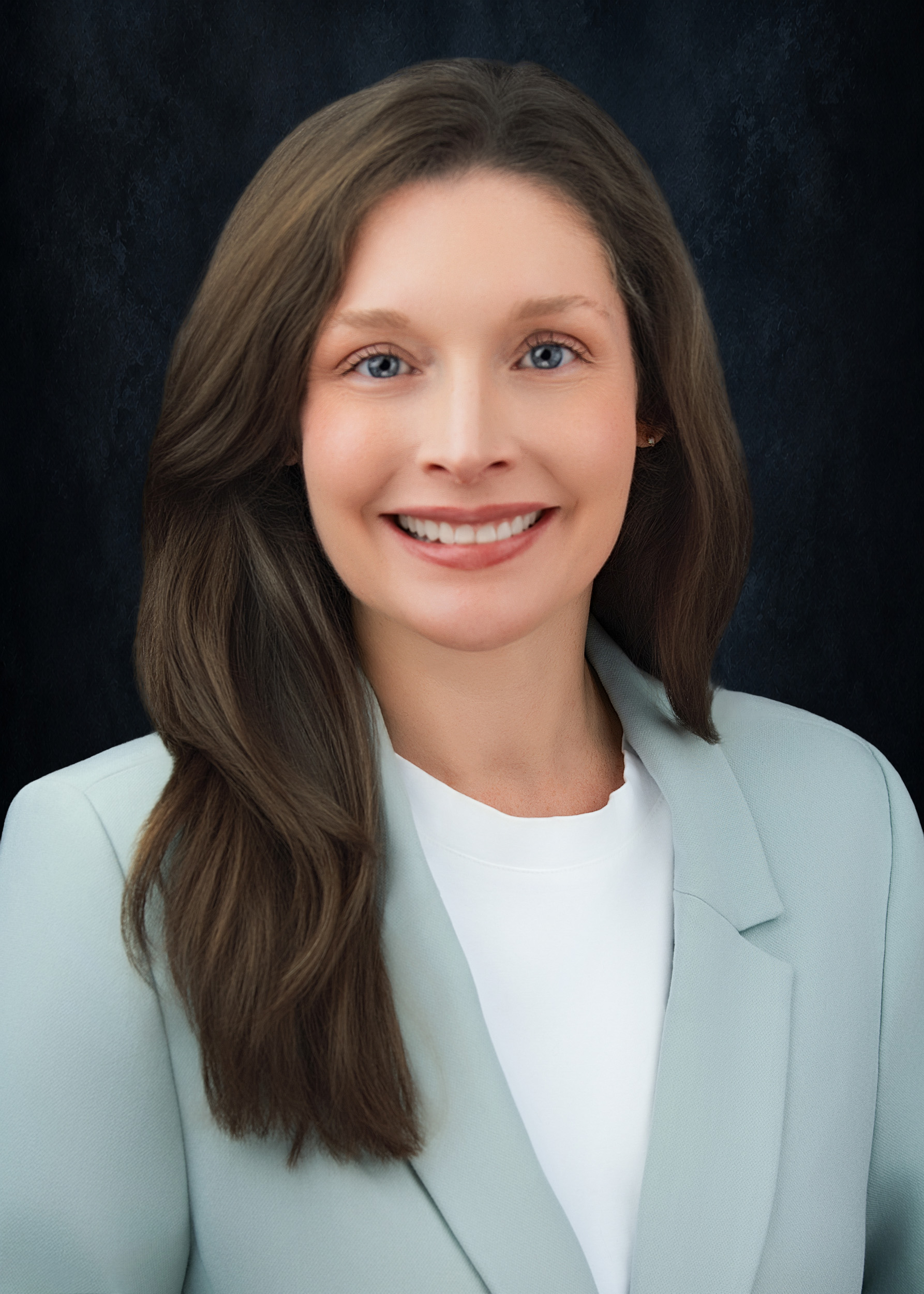 Professional headshot of a woman with long wavy dark brown hair and blue eyes, wearing a light sage green blazer over a white top, smiling warmly against a textured dark navy studio background.