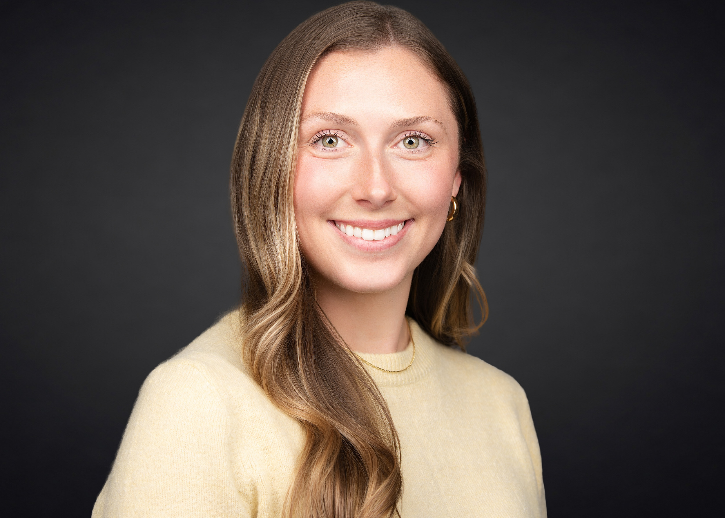 Professional headshot of a young woman with long wavy blonde-highlighted hair and green eyes, wearing a soft yellow crewneck sweater and small gold hoop earrings, smiling broadly against a dark charcoal background.