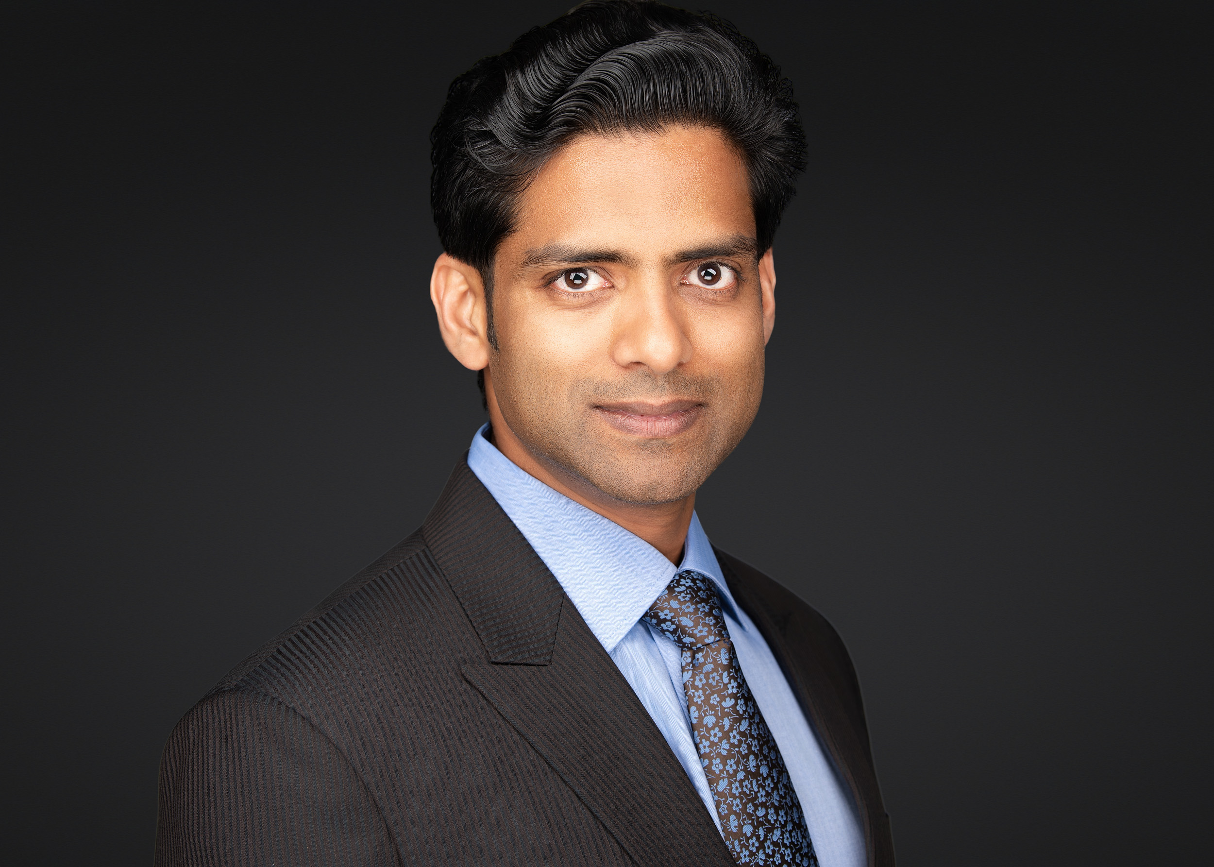 Professional headshot of a man with short dark hair, wearing a dark charcoal pinstripe suit, light blue dress shirt, and a patterned floral tie in brown and blue, with a composed expression against a dark black background.