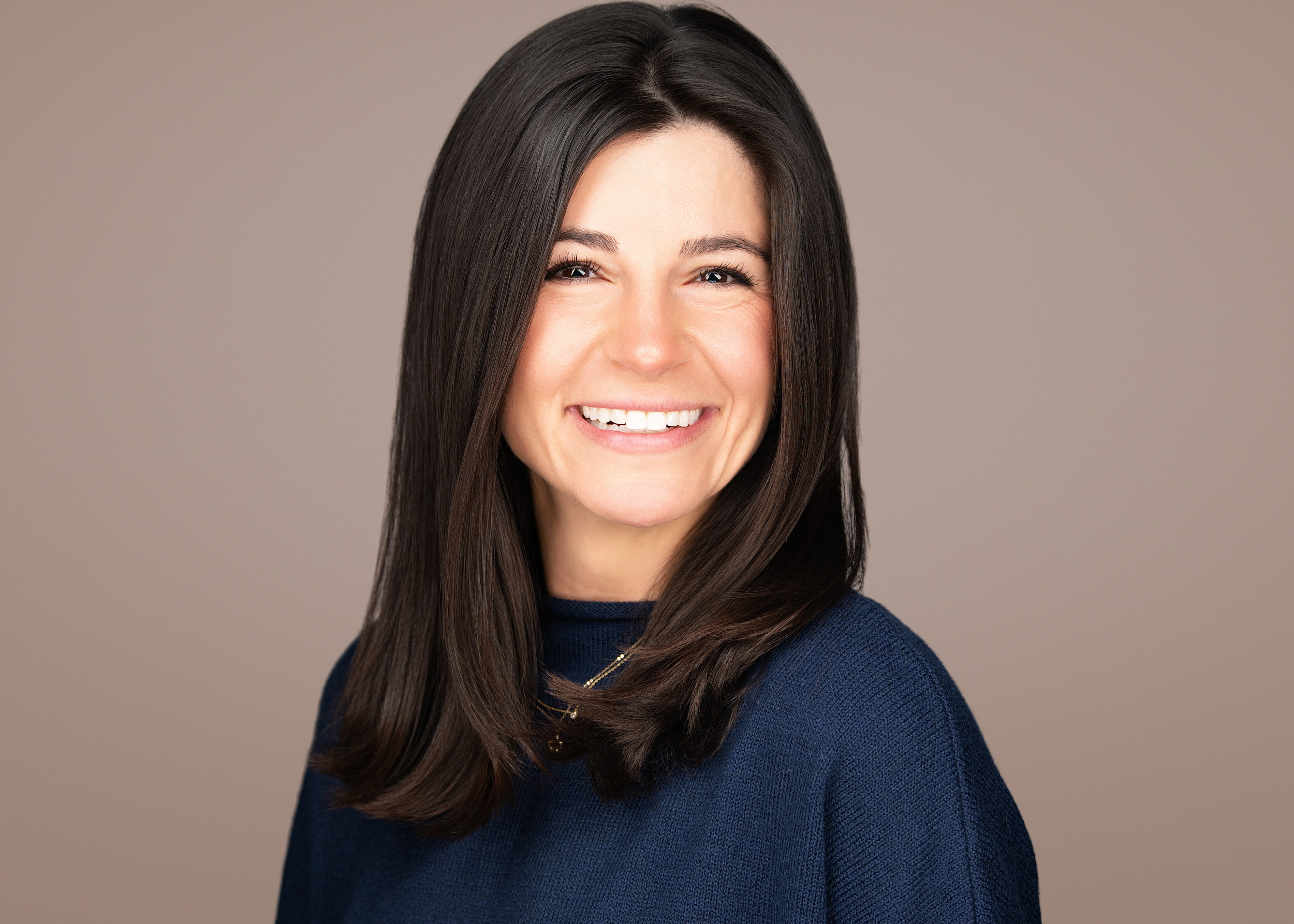 Professional headshot of a woman with long straight dark brown hair, wearing a navy turtleneck sweater and a delicate gold necklace, smiling warmly against a textured cream studio background.