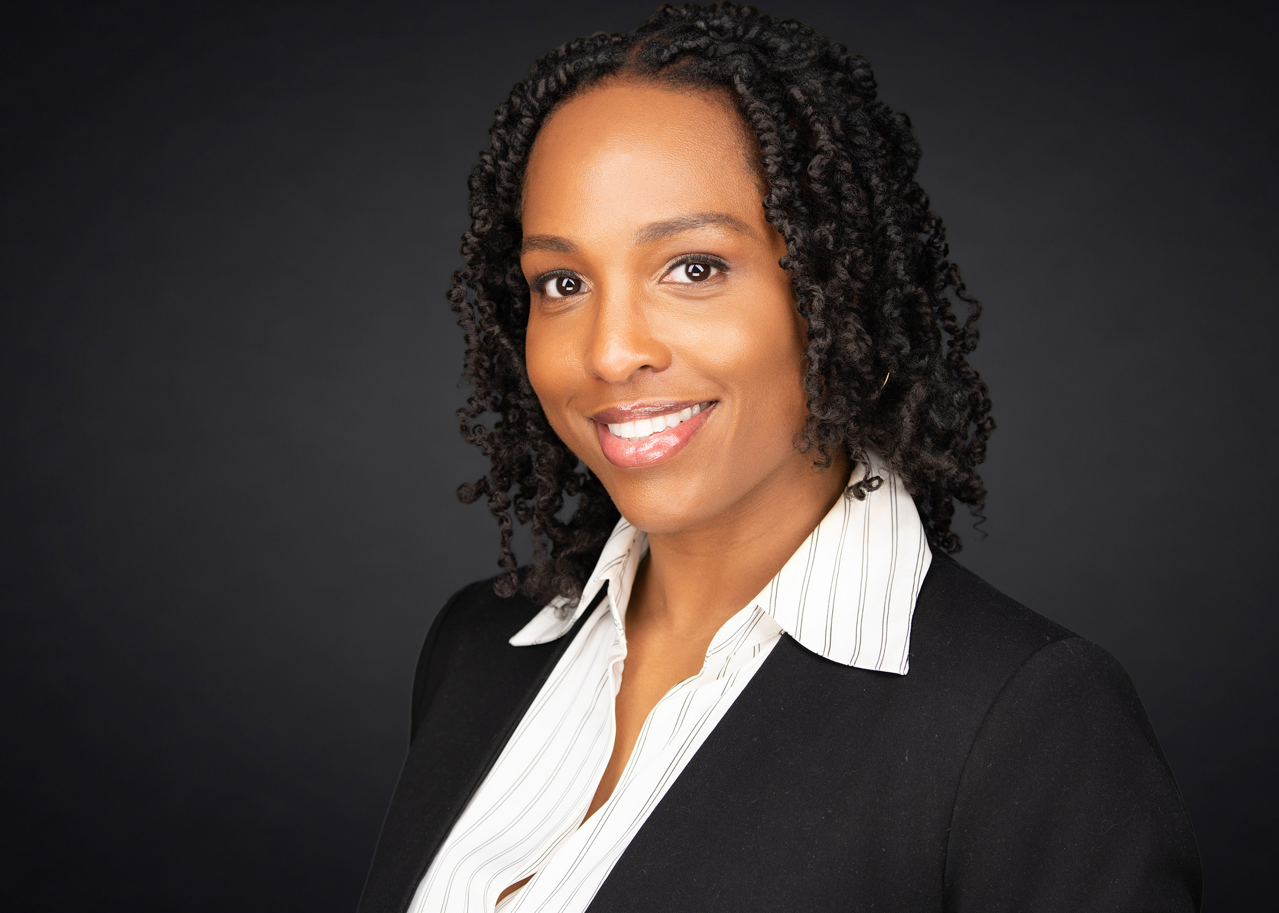 Professional headshot of a woman with natural twisted locs, wearing a black blazer over a white pinstripe shirt, smiling brightly against a dark charcoal studio background.
