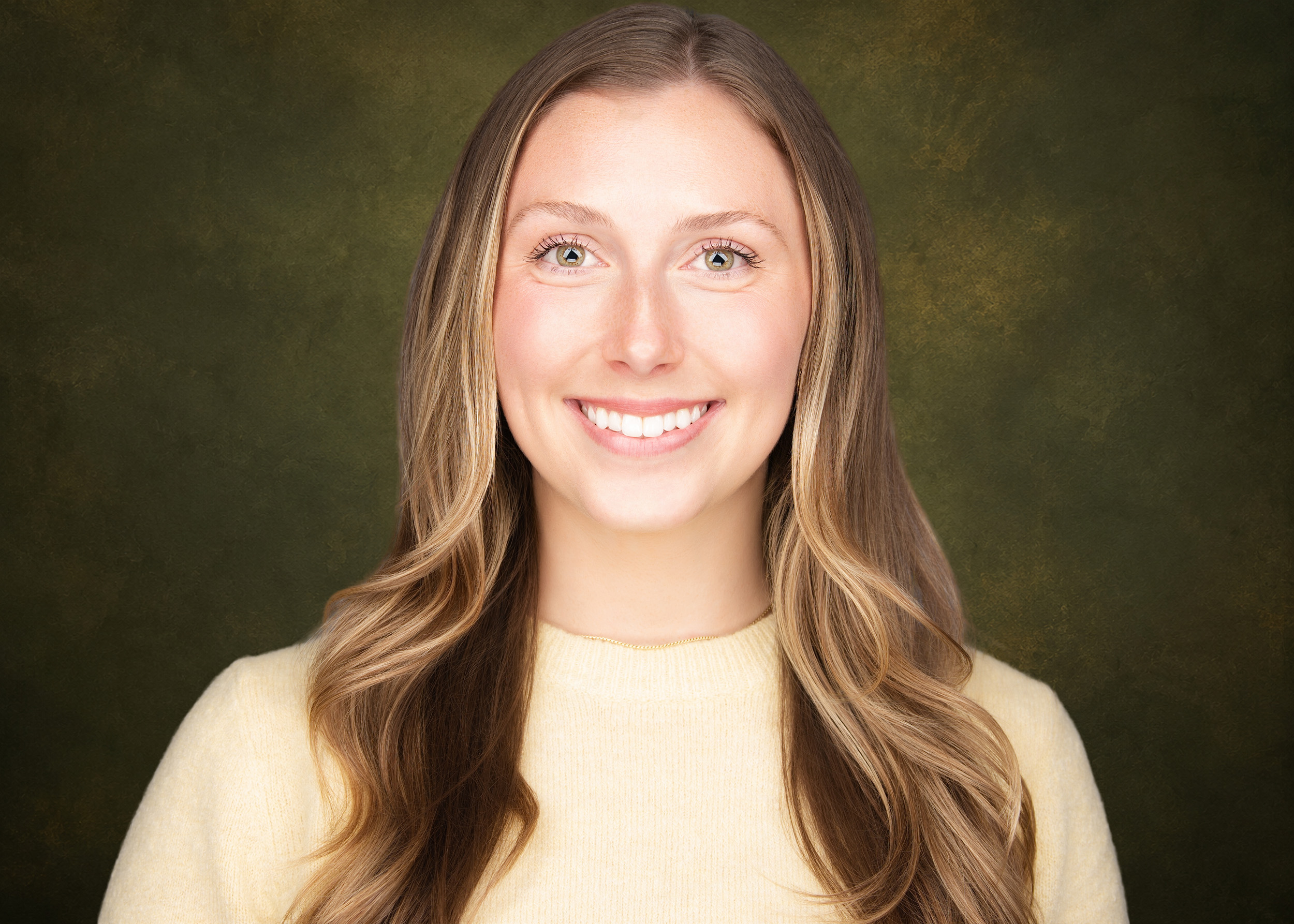 Professional headshot of a young woman with long wavy blonde-highlighted hair and green eyes, wearing a soft yellow crewneck sweater and small gold hoop earrings, smiling broadly against a dark charcoal background.